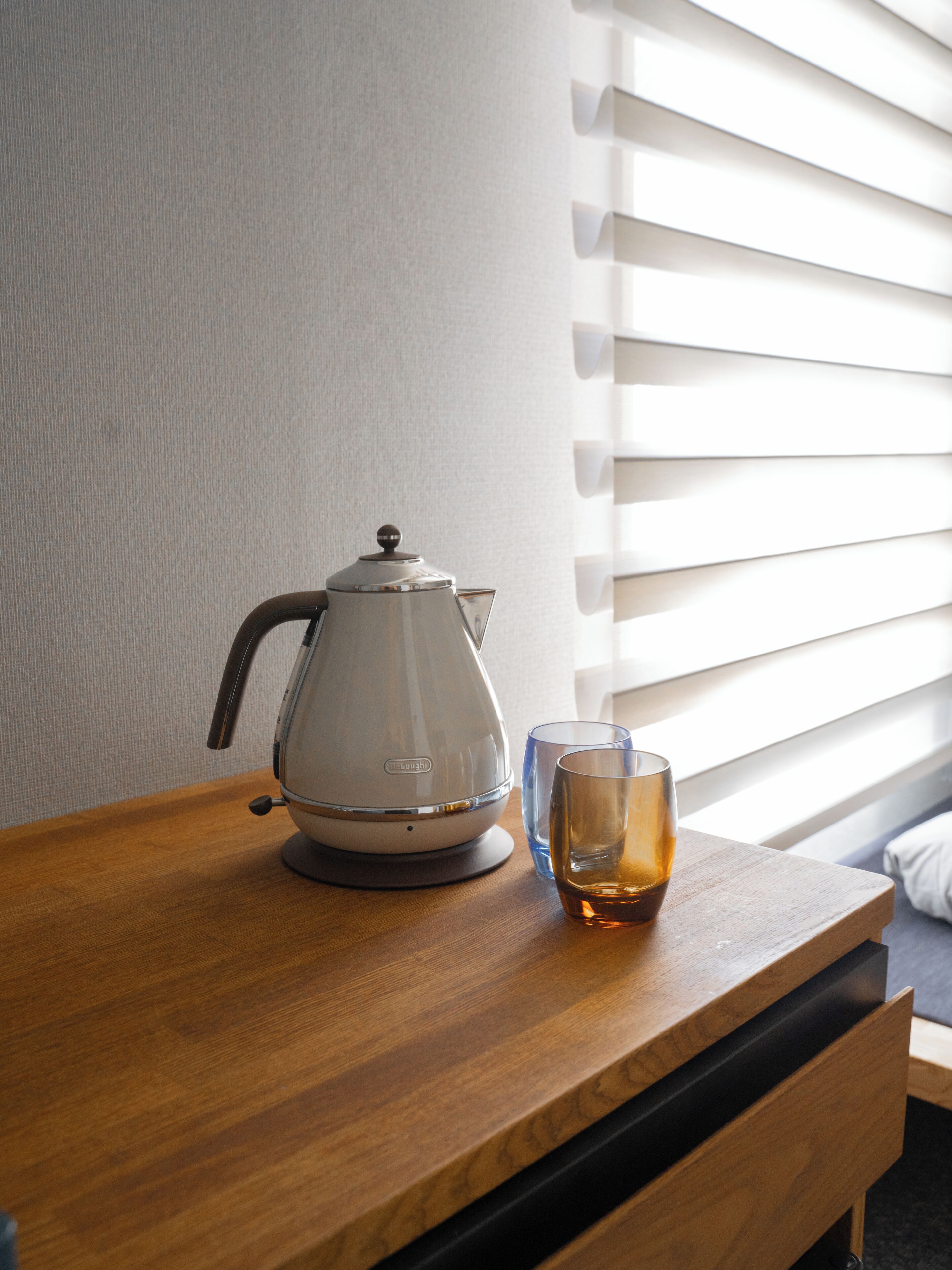 Una tetera eléctrica blanca sobre una mesa de madera, junto a un vaso de líquido ámbar. Se ve una ventana con persianas en el fondo.