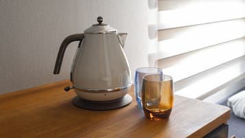 Una tetera eléctrica blanca sobre una mesa de madera, junto a un vaso de líquido ámbar. Se ve una ventana con persianas en el fondo.