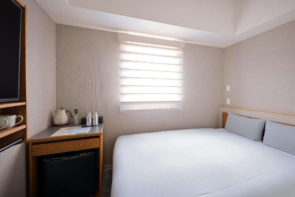 Habitación de hotel con cama, escritorio con cafetera y ventana con persianas.