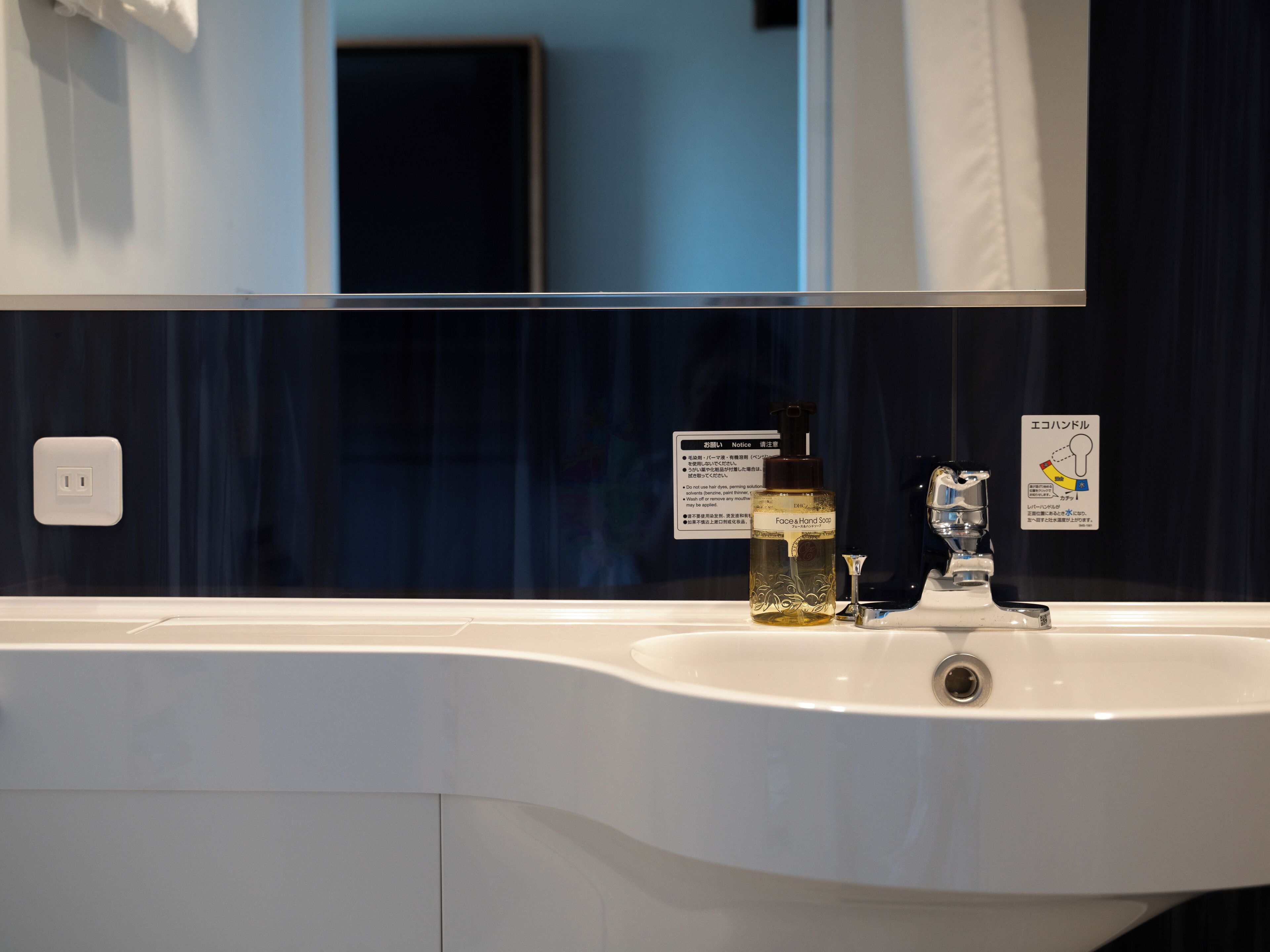 Un baño con un lavamanos blanco, un espejo y una pared azul oscuro. Hay un cartel con texto en japonés y una botella de jabón para manos.