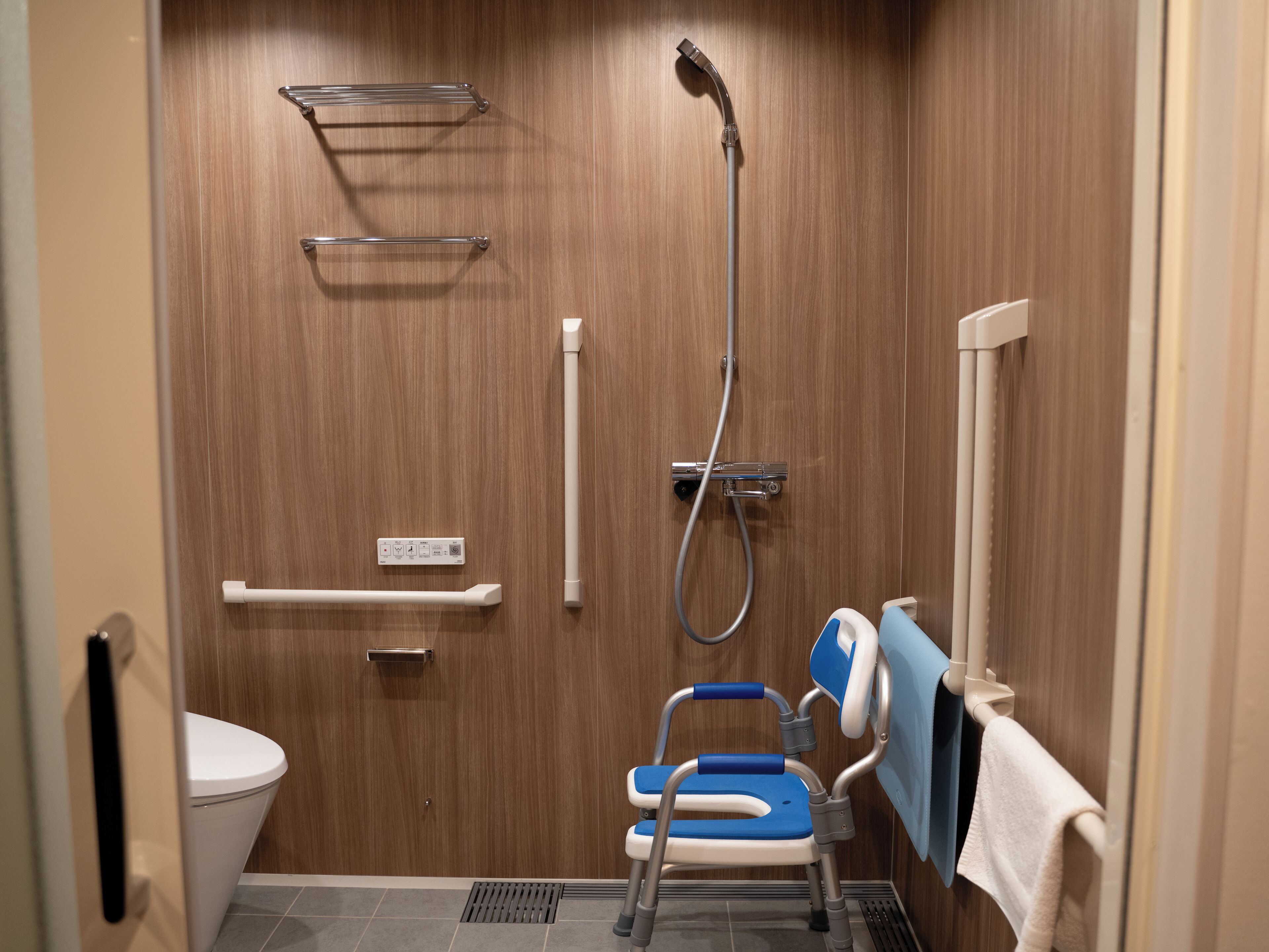 A small, compact bathroom with a wooden wall, a shower, a toilet, and a wheelchair-accessible bench.