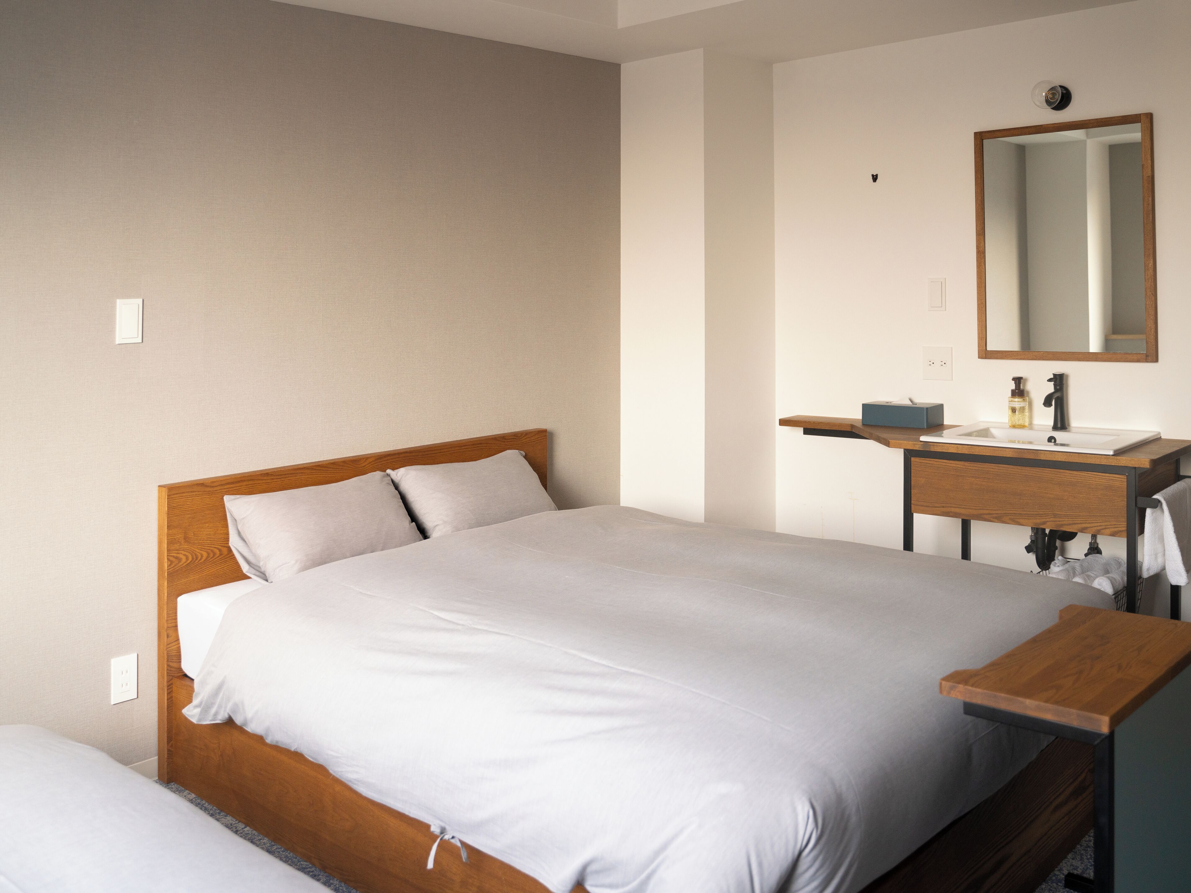A neatly made bed with a wooden headboard, a wooden bedside table, and a bathroom with a mirror, sink, and towel rack.
