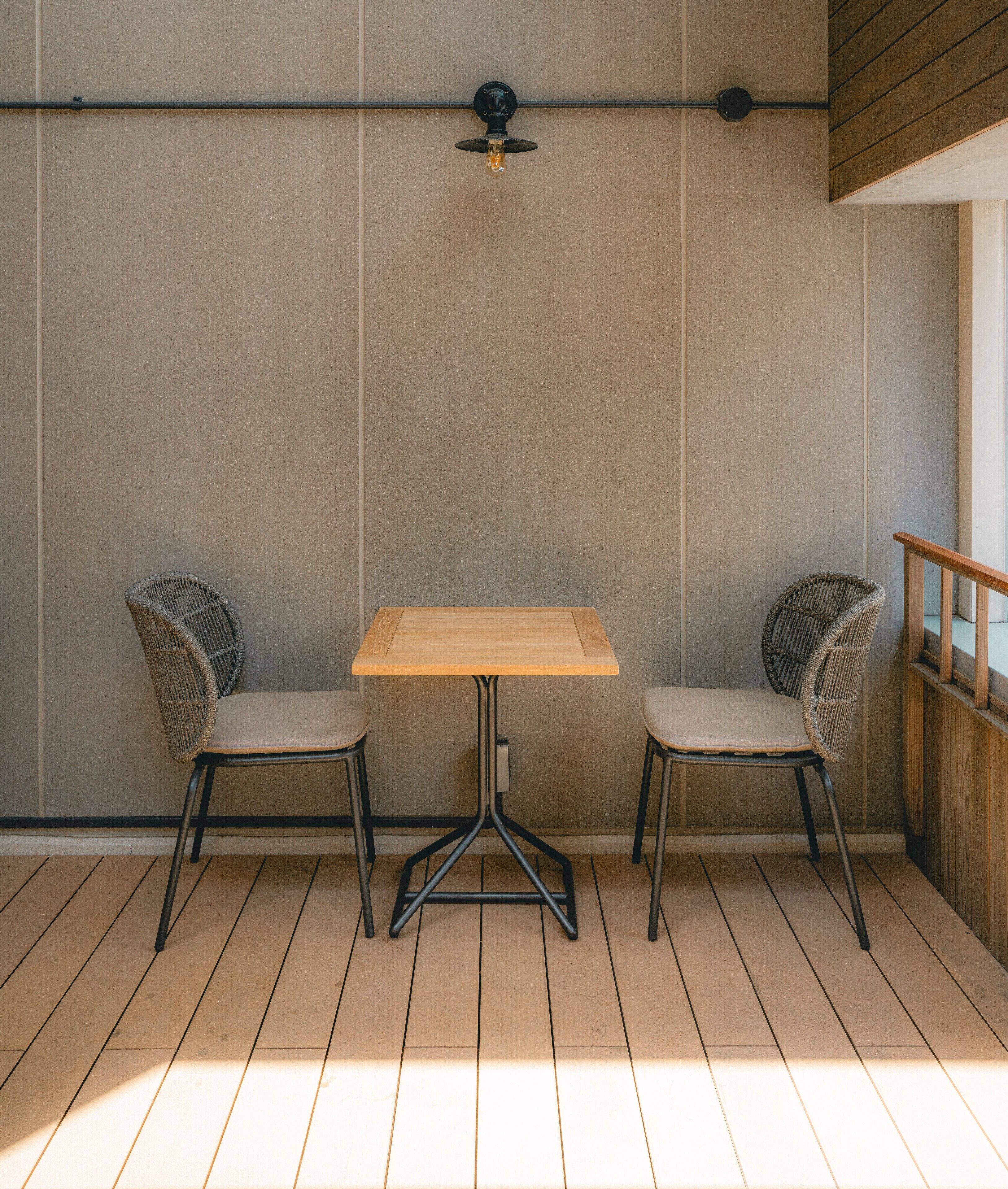 A minimalist room with a wooden table and two wicker chairs, a ceiling light fixture, and a wooden floor.