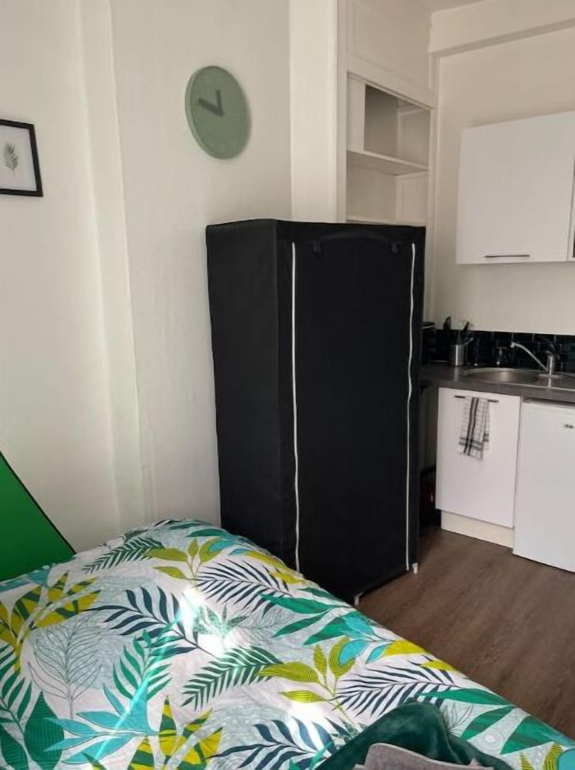 A compact room with a bed, a black refrigerator, a small kitchenette, and a green wall clock.