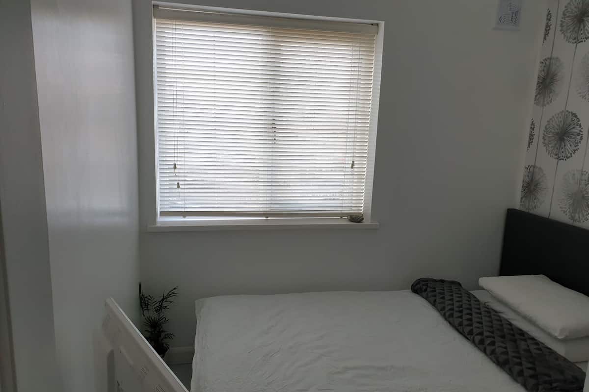 A small, neatly arranged bedroom with a single bed, a window with blinds, a radiator, and a wall with a floral pattern.