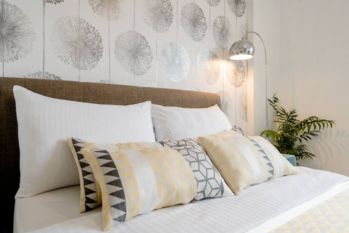 A neatly made bed with white and patterned pillows, a headboard, and a wall with a floral wallpaper design.
