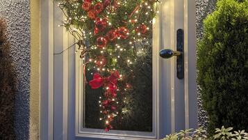 Una puerta principal con un adorno de corazón decorado con luces y adornos rojos, un buzón y una pequeña planta en maceta a su lado.