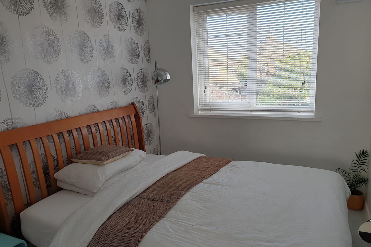 A neatly made bed with a wooden headboard, a window with blinds, and a patterned wallpaper.