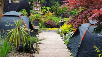 A pathway leading to two tents labeled "AmFlil" amidst a garden with various plants and a small birdhouse.