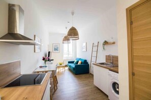 Cocina compacta con estufa, lavamanos y lavadora, zona de comedor con mesa y sillas, y un sofá azul en un espacio luminoso con un gran ventanal.