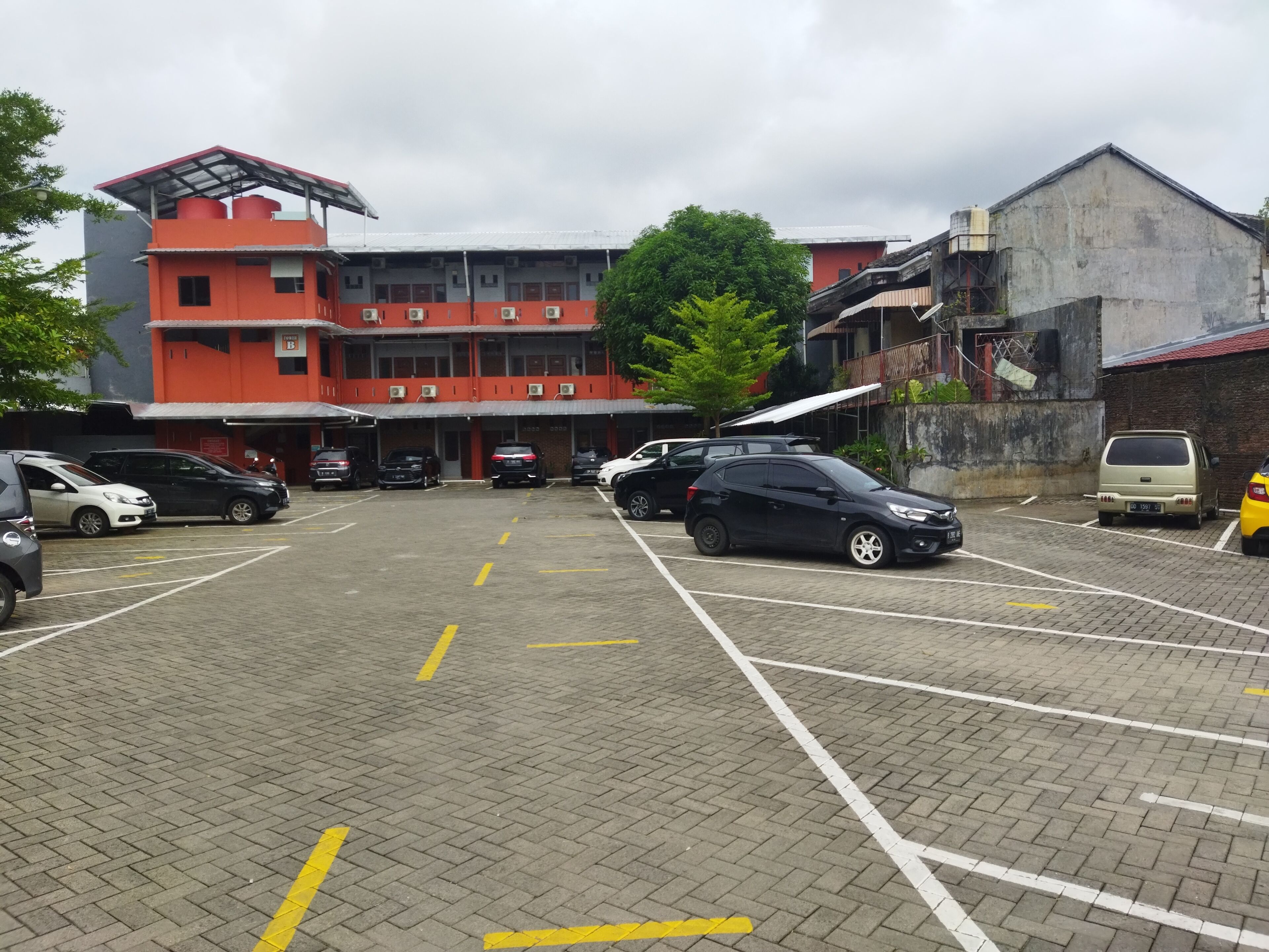 A parking lot with several cars, a building with red and white exterior, and a large tree in the background.