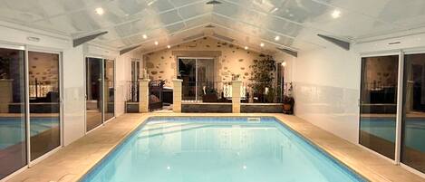 An indoor swimming pool with a tiled floor, glass doors on the sides, and a stone wall in the background.