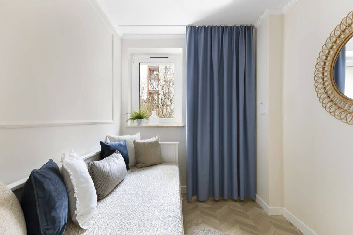 Quarto com cama, almofadas, janela com cortinas, espelho e uma planta no parapeito.