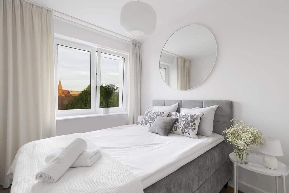 Quarto moderno com uma cama grande, um espelho redondo, mesa de cabeceira com luminária e janela com cortinas.
