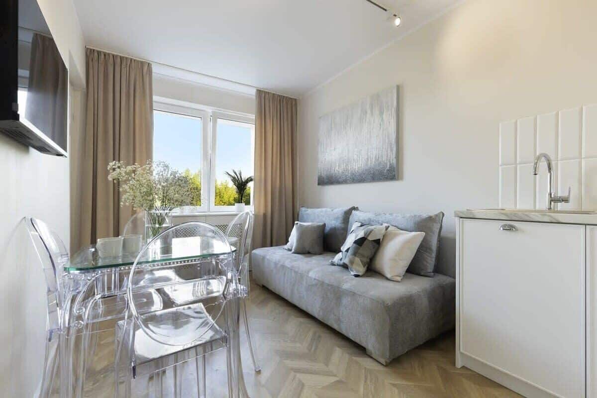 Uma sala de estar moderna com sofá cinza, mesa de jantar de vidro transparente e armários brancos.