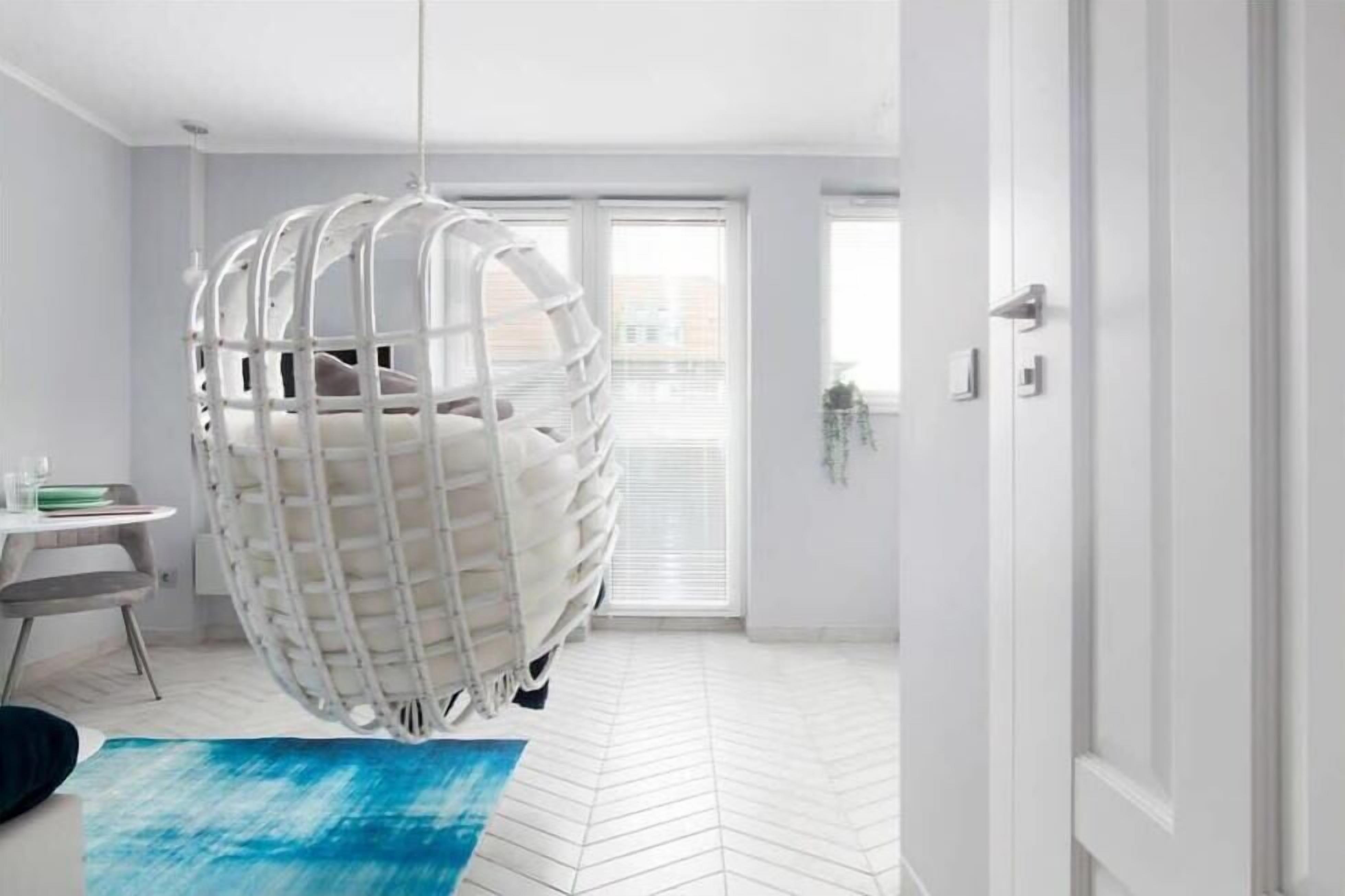 A room with a hanging chair, a small table with a chair, and a view of the outside through a window with blinds.