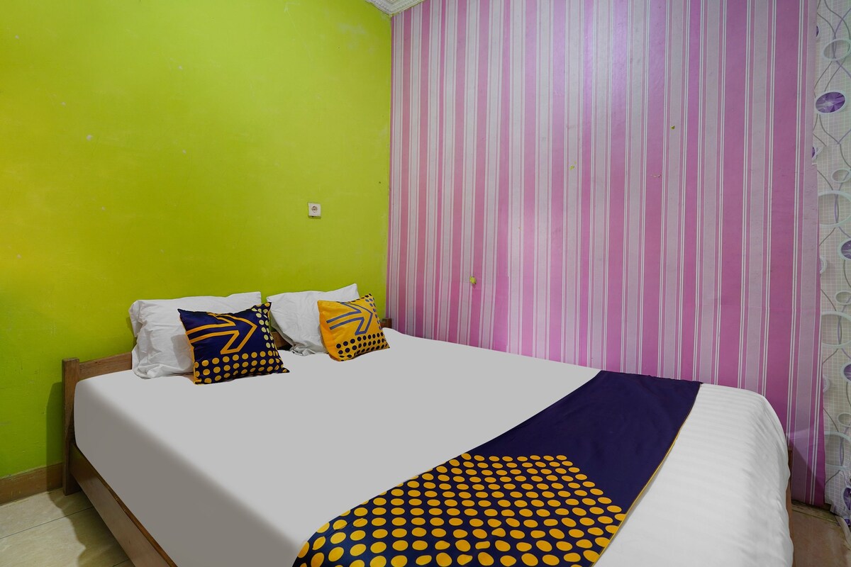 A bed with a white mattress, a blue and yellow patterned blanket, and two pillows with geometric designs. The room has a green wall and a striped curtain.