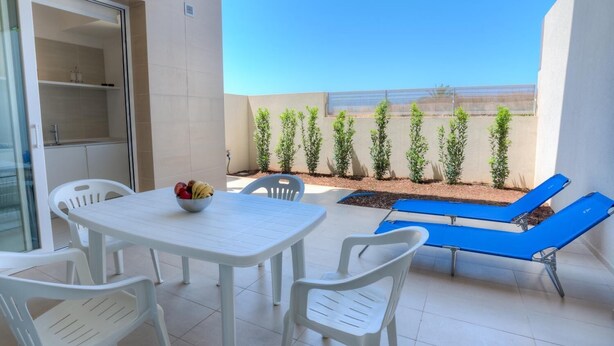 Un balcón con una mesa blanca, sillas, una silla reclinable azul y un cuenco de fruta. Hay plantas en macetas a lo largo de la pared.