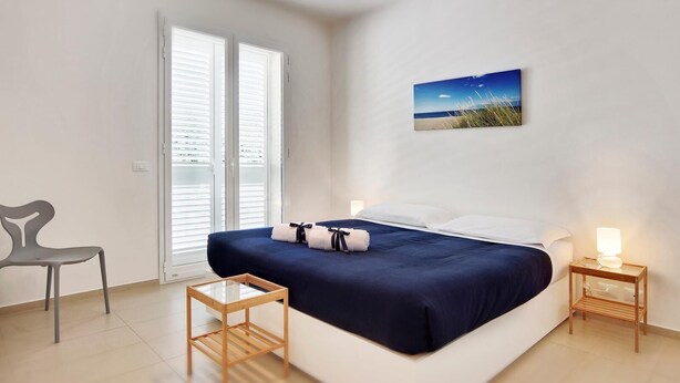 A bedroom with a bed, a chair, a bedside table, a picture on the wall, and a window with blinds. - Amazing Seaview Apartment with two bedroom (RAGUSA)