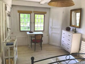 A room with a desk, chair, and a dresser. There are two windows with curtains, a door, and a hanging light fixture.