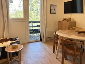 Una habitación con una mesa de comedor de madera, sillas, una mesita redonda con una cesta, un sofá con una manta de estampado vaca, un televisor en la pared y una puerta que da a un espacio exterior con una mesa y sillas.