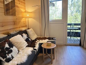 A cozy room with a wooden headboard, a sofa with a cowhide cover, a small round table with a lamp, and a view of a deck with outdoor furniture. - Les Orres 1650: Cozy studio 4 ppl, shuttle, washer (Les Orres)