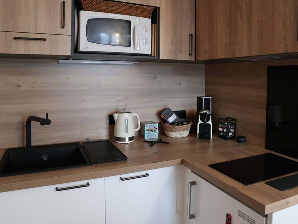 Una cocina moderna con armarios de madera, un lavamanos de acero inoxidable, un microondas, un hervidor, una cafetera y una placa de inducción negra.