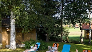 A garden with four lounge chairs under trees, a wooden structure, and a red-roofed building in the background. - Pension Wiesengrund (Neuschoenau)