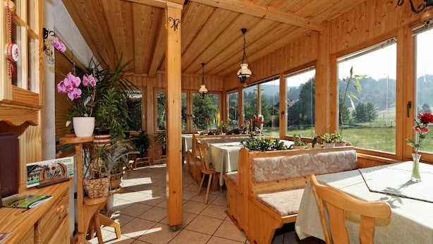 A wooden interior with tables and benches, large windows, and potted plants. - Pension Wiesengrund (Neuschoenau)