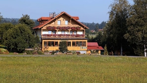 A two-story wooden building with a balcony, surrounded by a field and trees. - Pension Wiesengrund (Neuschoenau)