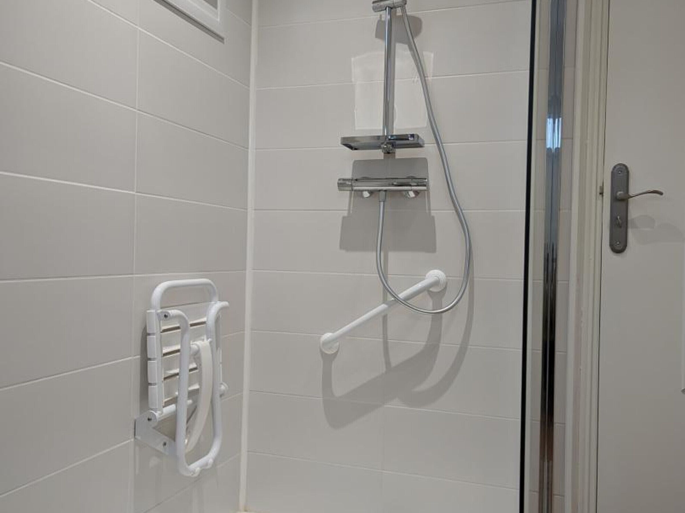 Een moderne douche met een handdouche en een handkraan, een glazen deur en een witte betegelde wand.