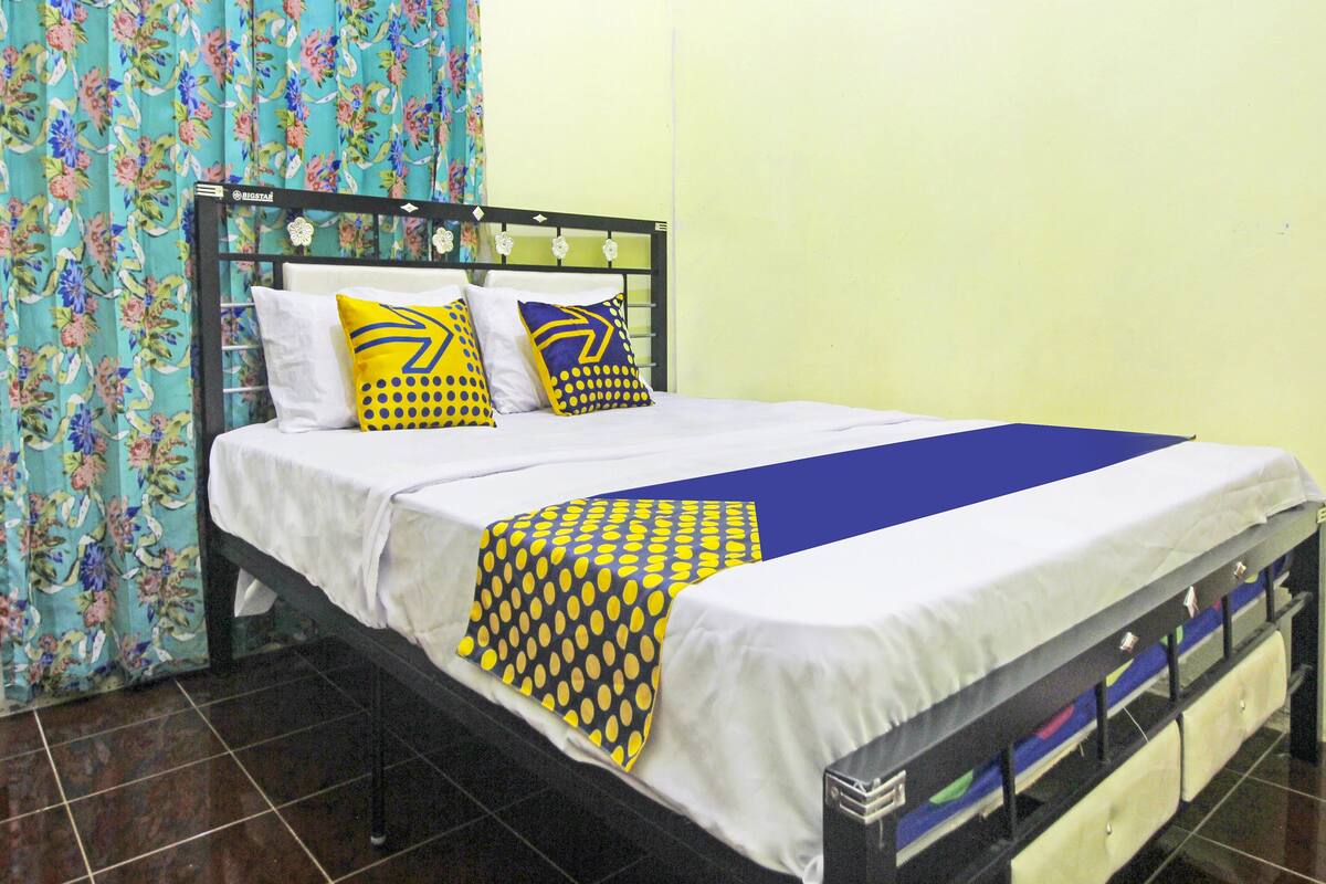 Un lit recouvert d’une couvre-lit blanche et bleue, deux oreillers à motifs jaunes et bleus, et un rideau à fleurs.
