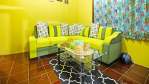A living room with a bright yellow wall, a green sofa with patterned cushions, a glass coffee table with decorative items, and a floral curtain. - Hotel O Desa Wisata NangelasariNearPasir Tulang (Nangelasari)