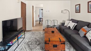 Ein Wohnzimmer mit Fernseher, einer Couch und einer Holzkiste. Im Hintergrund stehen Stühle und ein Tisch.