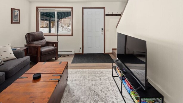 Ein Wohnzimmer mit braunem Ledersessel, grauem Sofa und einem Holztisch. An der Wand befindet sich ein Fernseher auf einem Schrank, und darüber ein Flur mit Kleiderhaken.