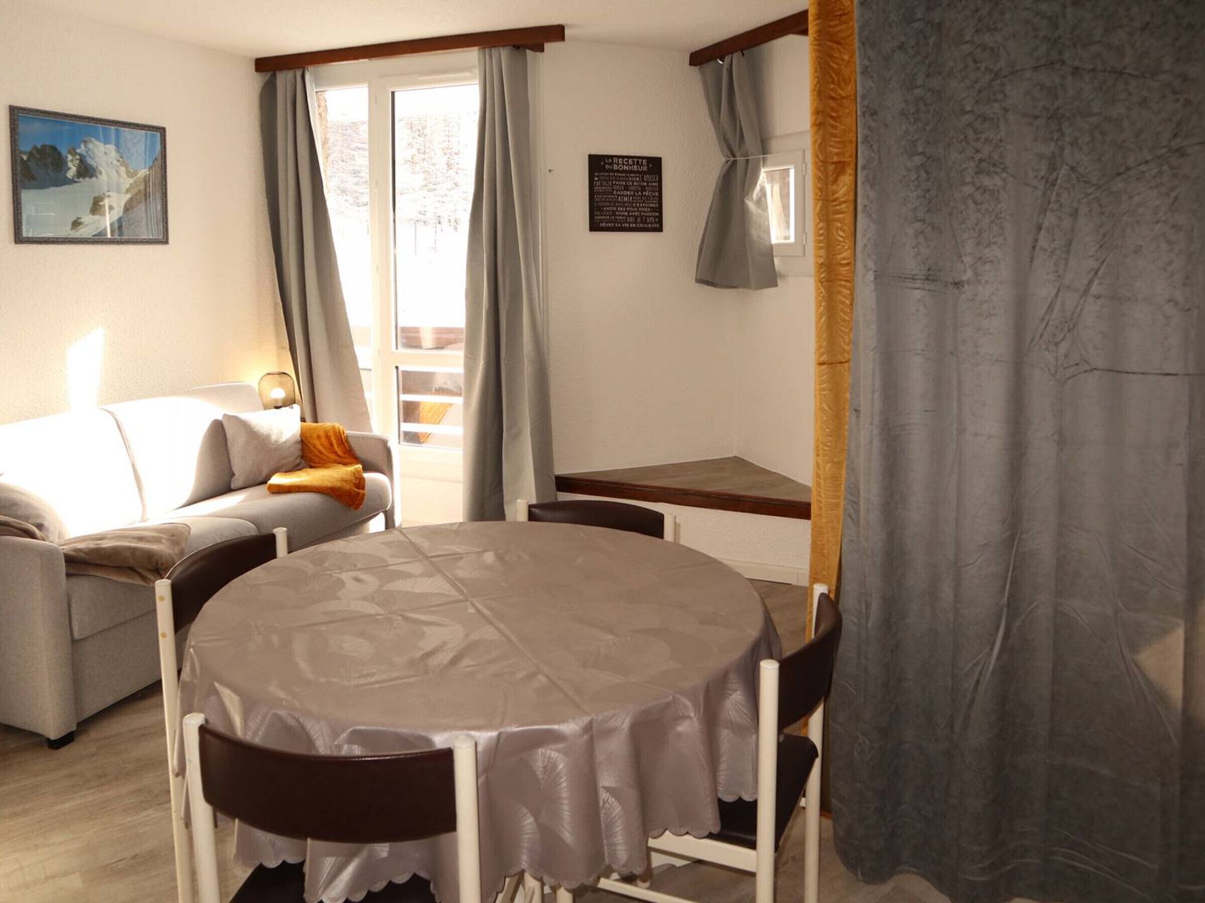 A cozy room with a round table, a sofa, and a chair. There is a window with curtains, a painting on the wall, and a sign with text.