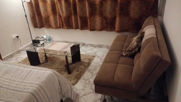 A small, furnished room with a sofa, a glass coffee table, a bed with a striped blanket, and a patterned curtain. - Lovely spacious and quiet small budget Room is located in the Center of AbuDhabi (AbuDhabi)