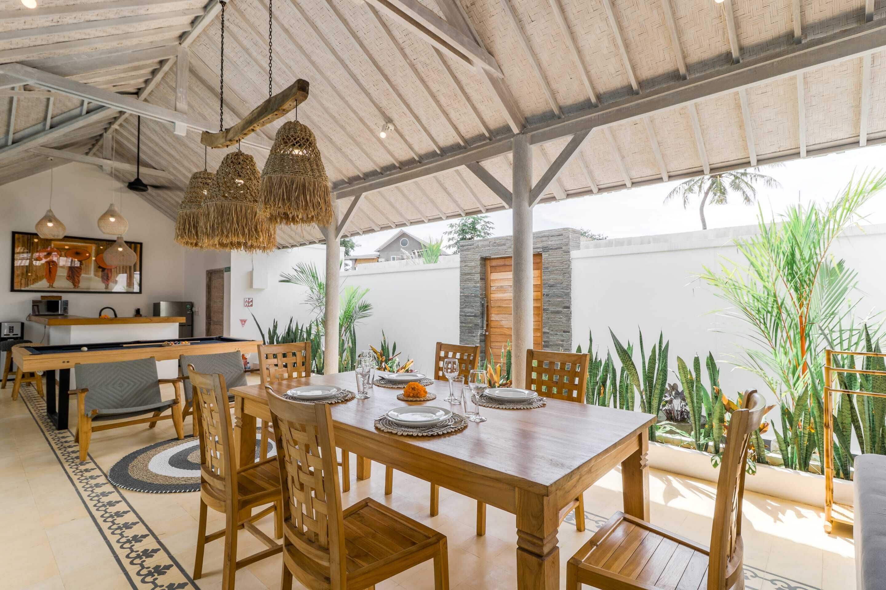 A dining area with a wooden table set for four, surrounded by chairs, under a thatched roof.