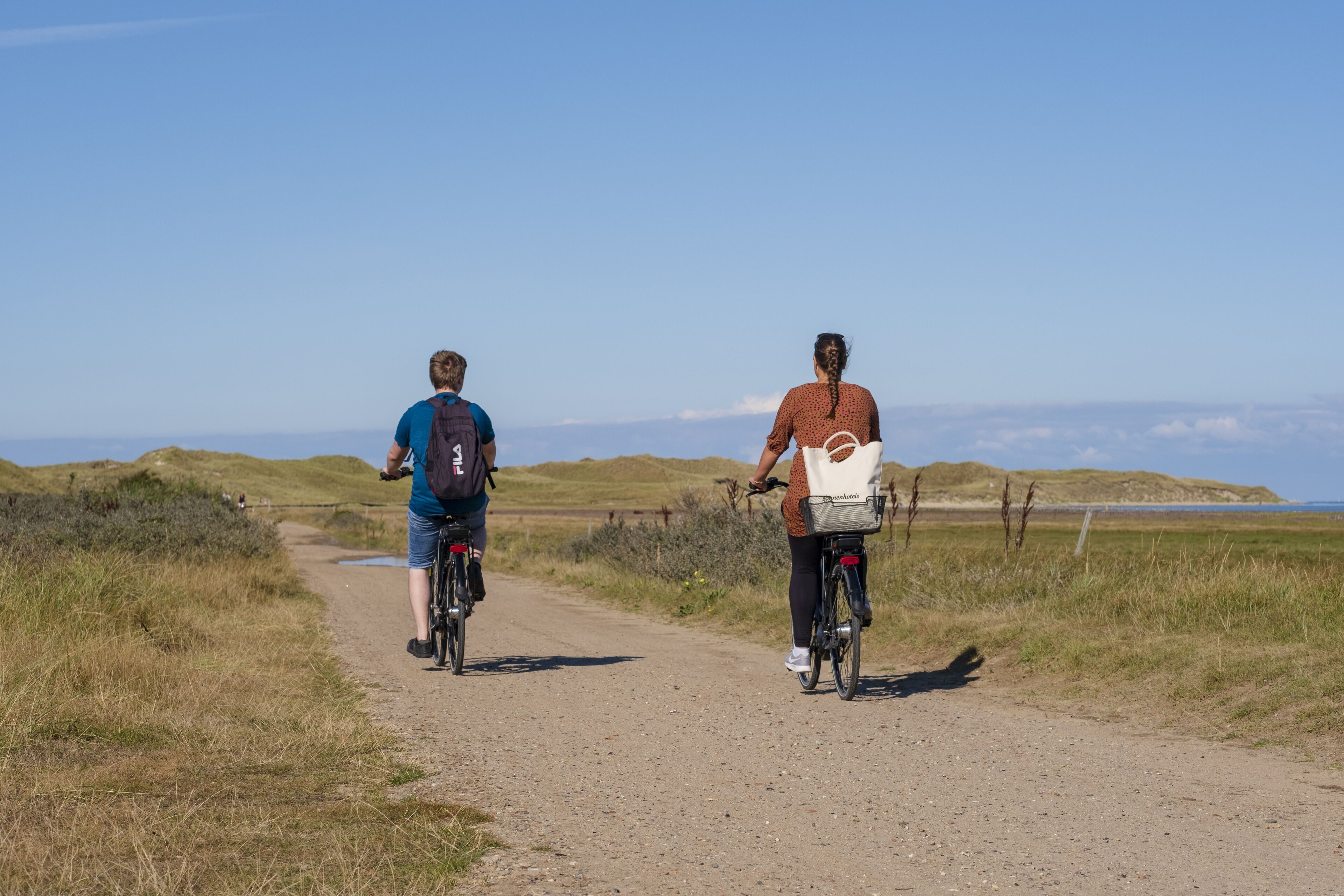 Two people riding bicycles on a dirt path with grassy hills in the background.