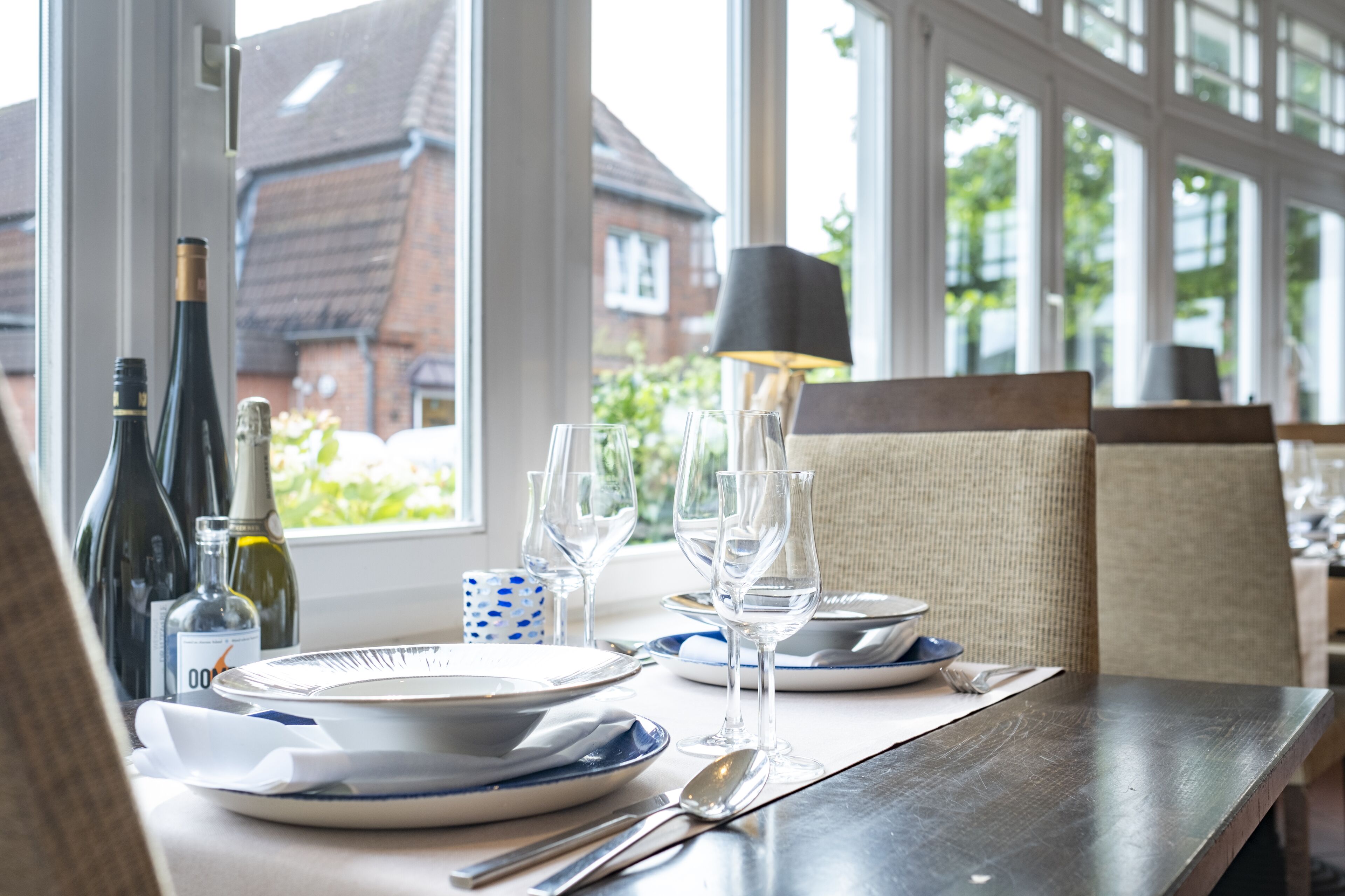 A dining table set with plates, glasses, and cutlery, with wine bottles and a lamp on the windowsill.