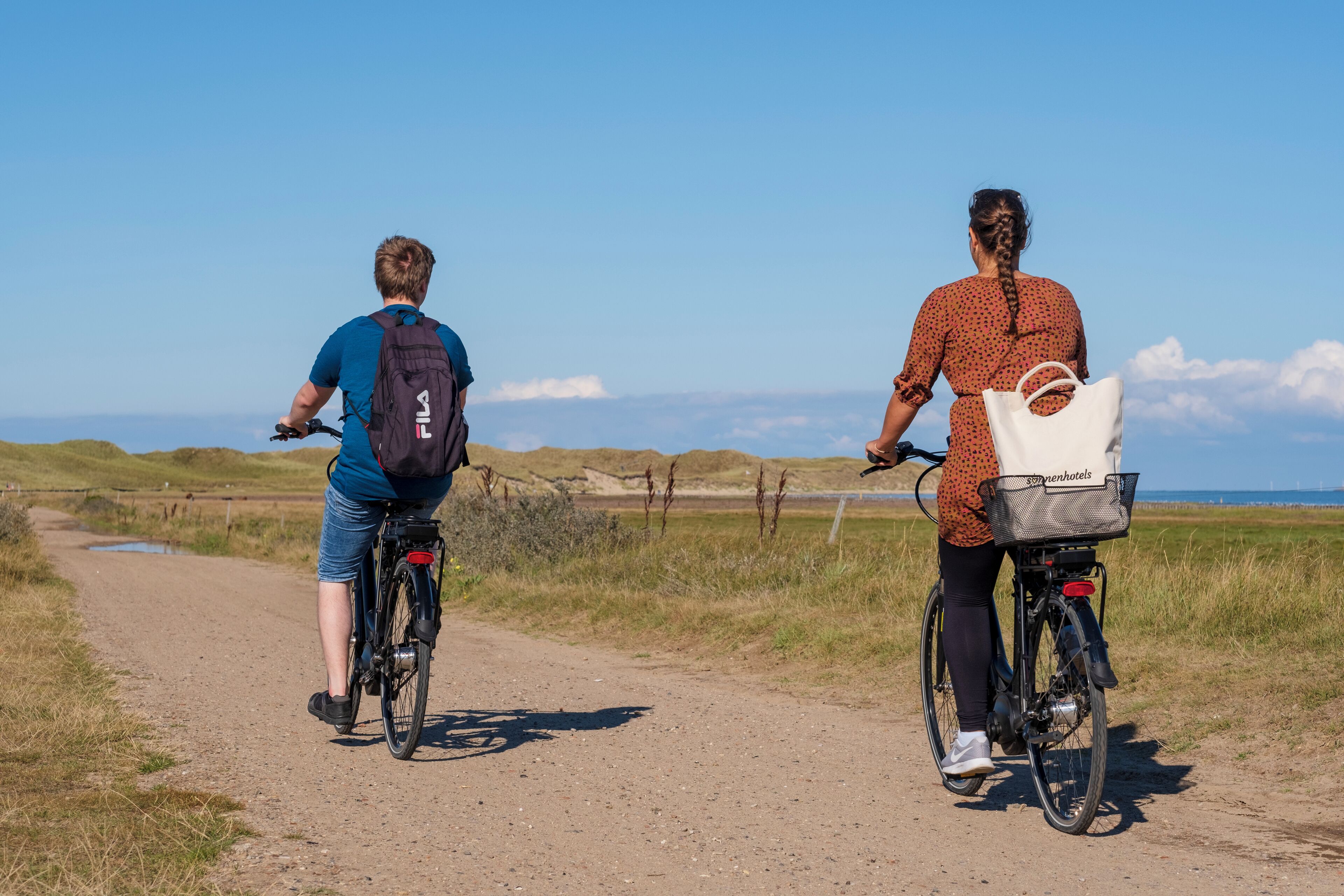 Two people riding bicycles on a dirt path with a scenic view of the sea and hills in the background.