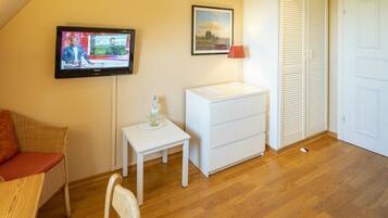 A room with a television mounted on the wall, a small white table with a bottle, a white drawer unit, a wicker chair, and a wooden floor.