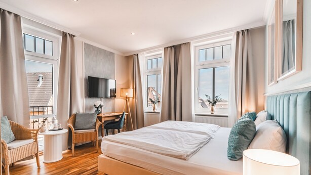 Una habitación de hotel con una cama grande, dos sillas, un escritorio, un televisor y ventanas amplias.