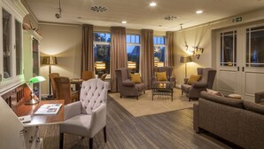 Un salón amplio y bien iluminado con asientos cómodos, incluyendo sillones y sofás, una mesa de centro de madera y un escritorio con lámpara. Grandes ventanas con cortinas permiten luz natural y vistas.