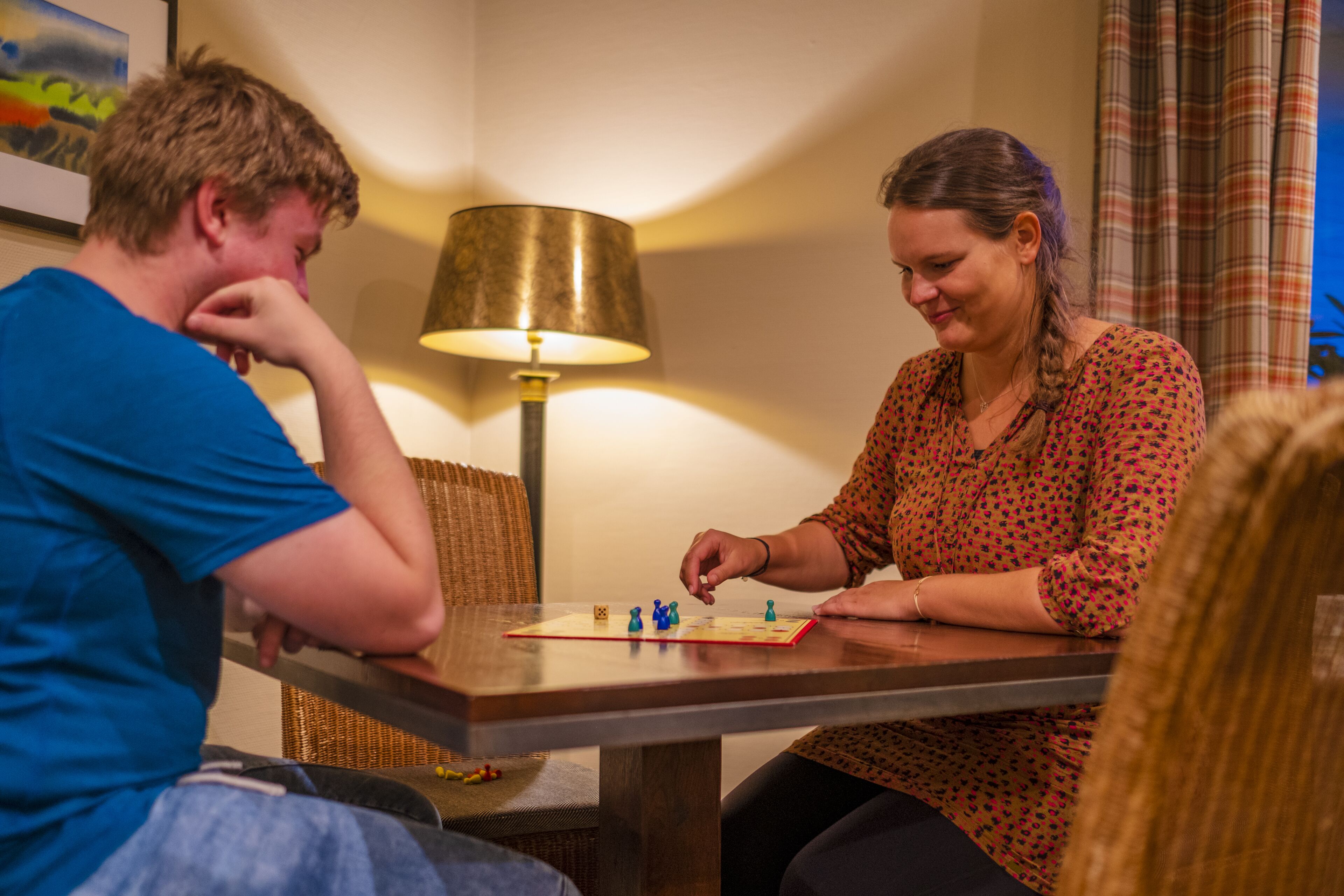 Two people playing a board game at a wooden table with a lamp in the background.