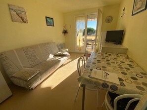 A living room with a sofa, a dining table with chairs, a television, and a window with curtains. - Charming T2 Apartment near Pontaillac Beach with Pool (Vaux-sur-Mer)