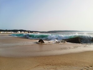 A beach with waves, a rock formation, and buildings in the background.