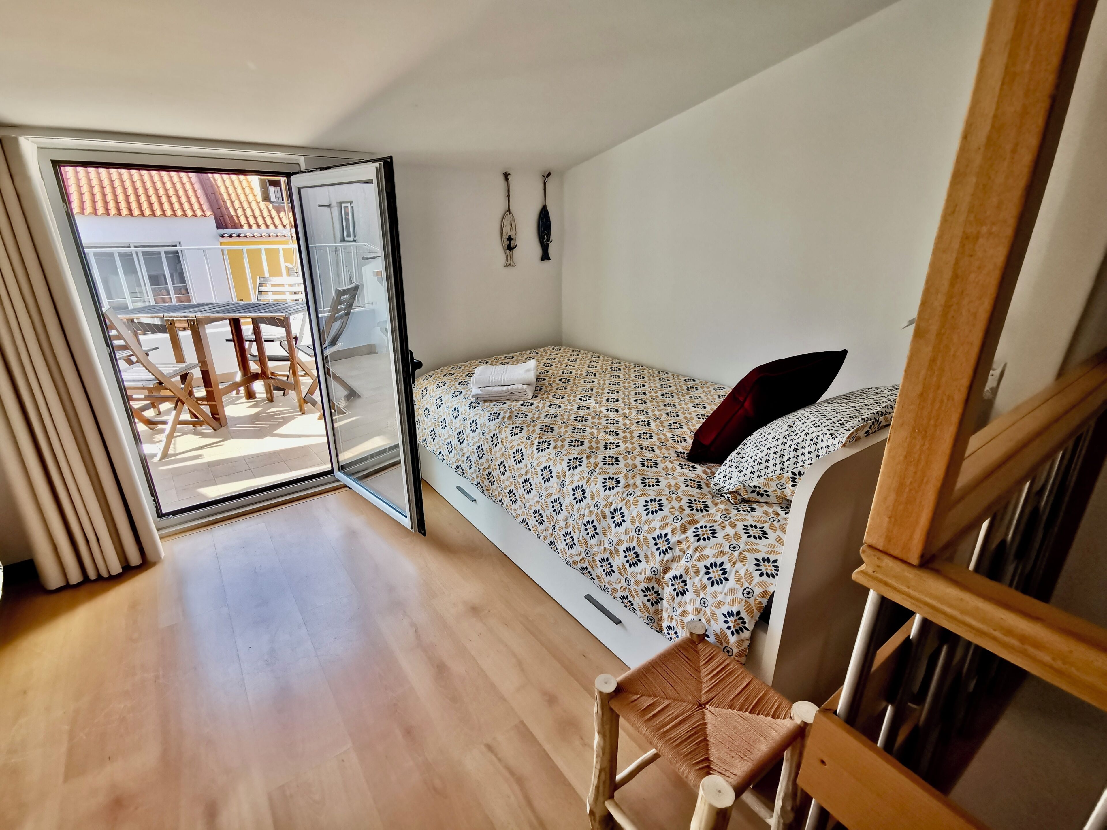 A room with a bed, a wooden chair, a table, and a sliding glass door leading to an outdoor area with a table and chairs.