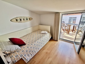A single bed with a patterned bedspread, a red pillow, a wall-mounted light fixture, and a sliding glass door leading to an outdoor seating area.