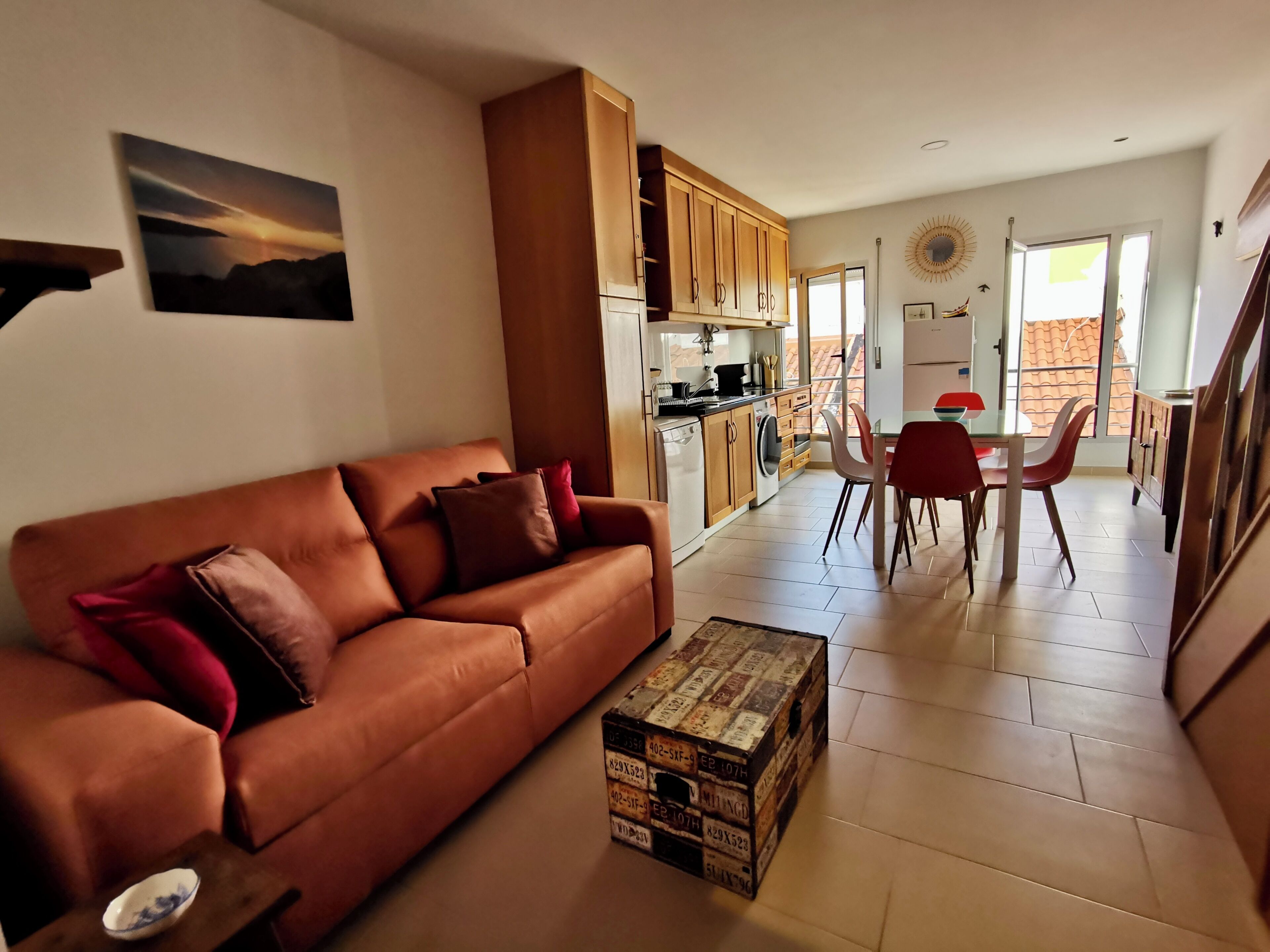 A living room with a brown sofa, a dining area with a table and chairs, a kitchen with wooden cabinets, and a balcony with a view.